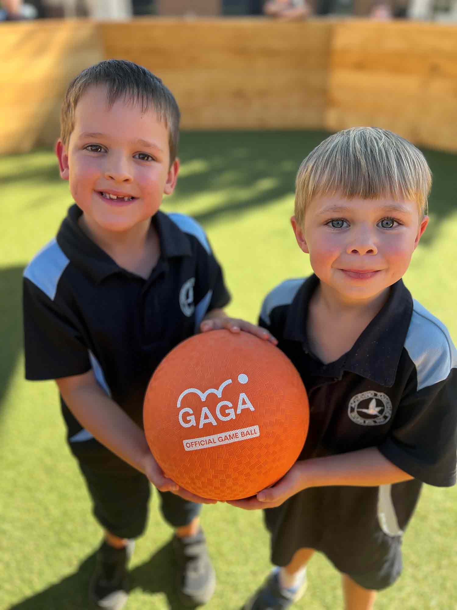 Two primary school children in school uniform inside a Gaga Ball Pit - Proudly holding an orange Gaga Ball.