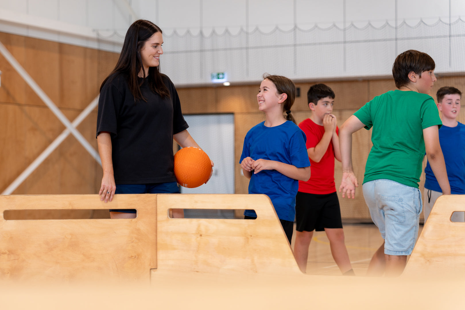 A PE teacher holding an orange gaga ball smiles at a group of primary school students standing inside a wooden gaga ball pit in a school gymnasium.